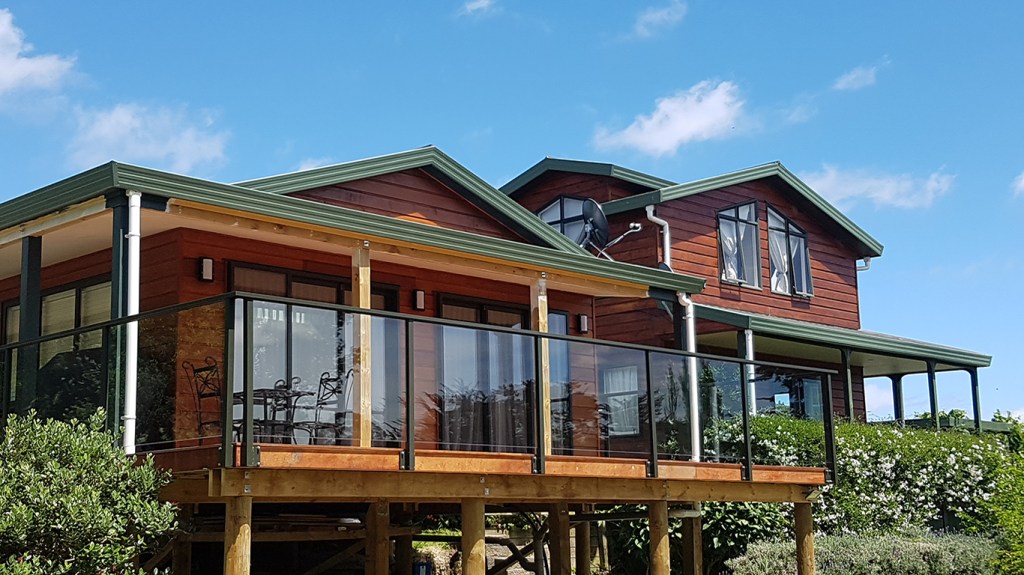 A modern house featuring a wooden deck with a frameless glass balustrade, surrounded by greenery and under a blue sky.