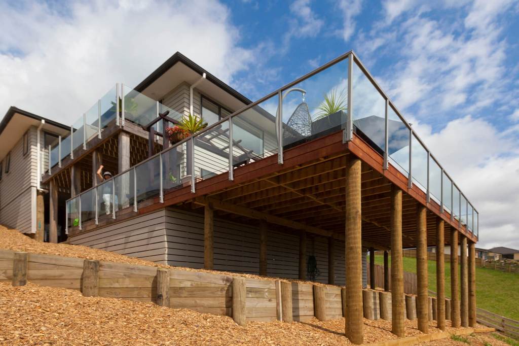 A modern house with a spacious balcony featuring frameless glass balustrades, set on stilts, overlooking a hillside.