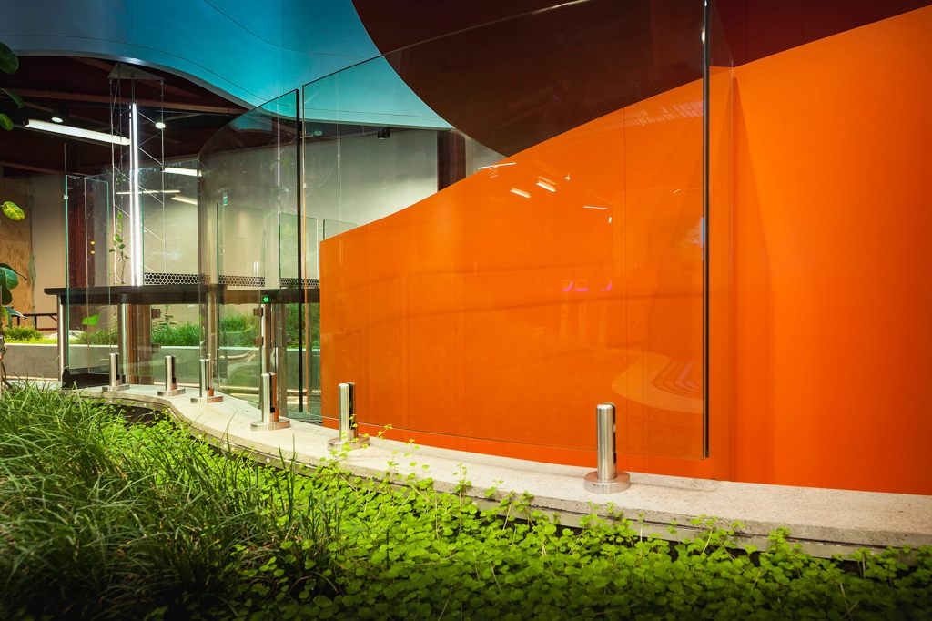 Frameless glass balustrade with stainless steel supports, featuring a vibrant orange wall and lush greenery in an indoor setting.