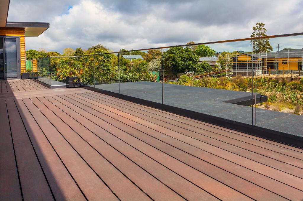 Outdoor area featuring a modern wooden deck with frameless glass balustrades overlooking greenery and a residential area.