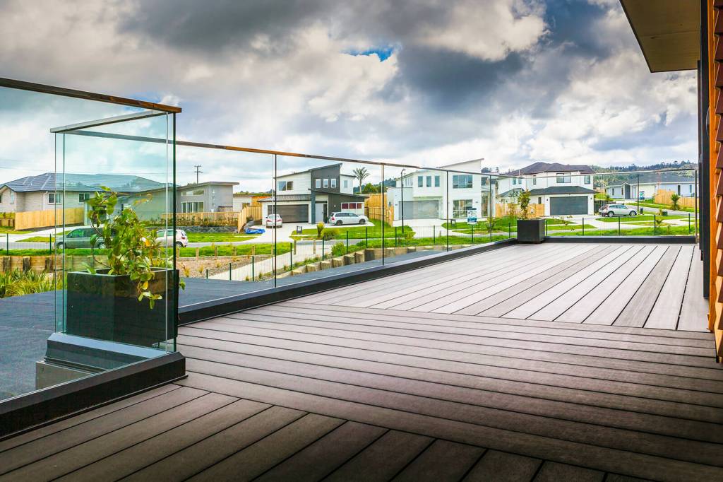 A spacious outdoor deck with a frameless glass balustrade overlooking a neighborhood, featuring wooden flooring and planters.