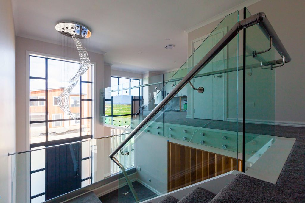 Interior view showcasing a glass balustrade with a stainless steel handrail, leading to a staircase. The space features large windows providing a bright atmosphere and a modern chandelier hanging from the ceiling.