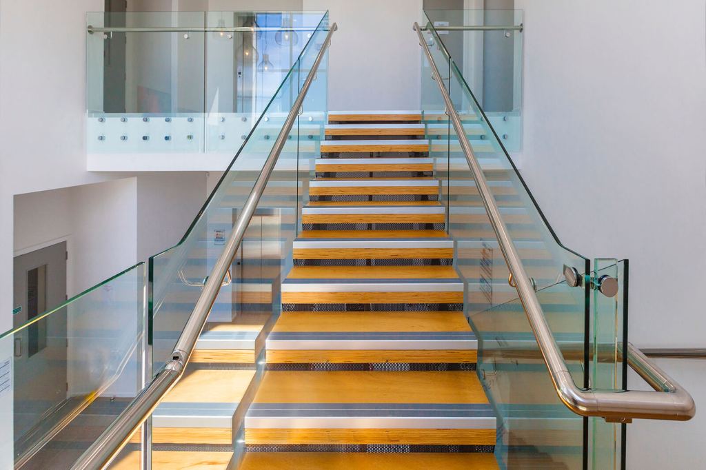 A modern staircase featuring frameless glass balustrades and a stainless steel handrail, showcasing a sleek design with wooden steps.