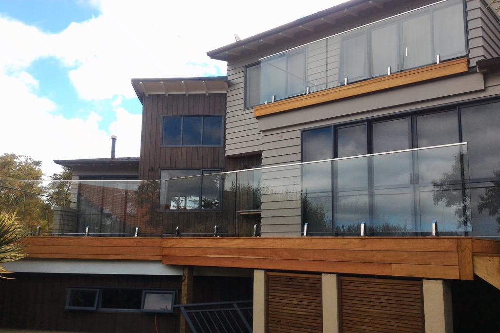 A modern residential building featuring a balcony with frameless glass balustrades, complemented by wooden decking and a mix of wooden and gray siding.