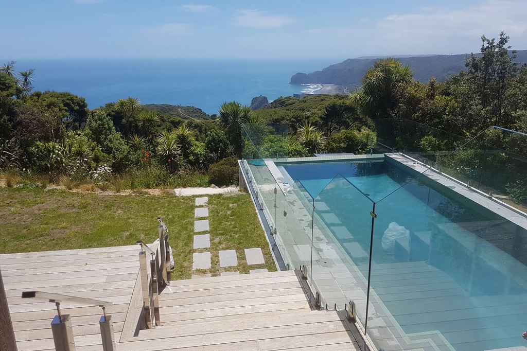 A picturesque view from a backyard featuring a glass balustrade surrounding a swimming pool, with lush greenery and ocean in the background.